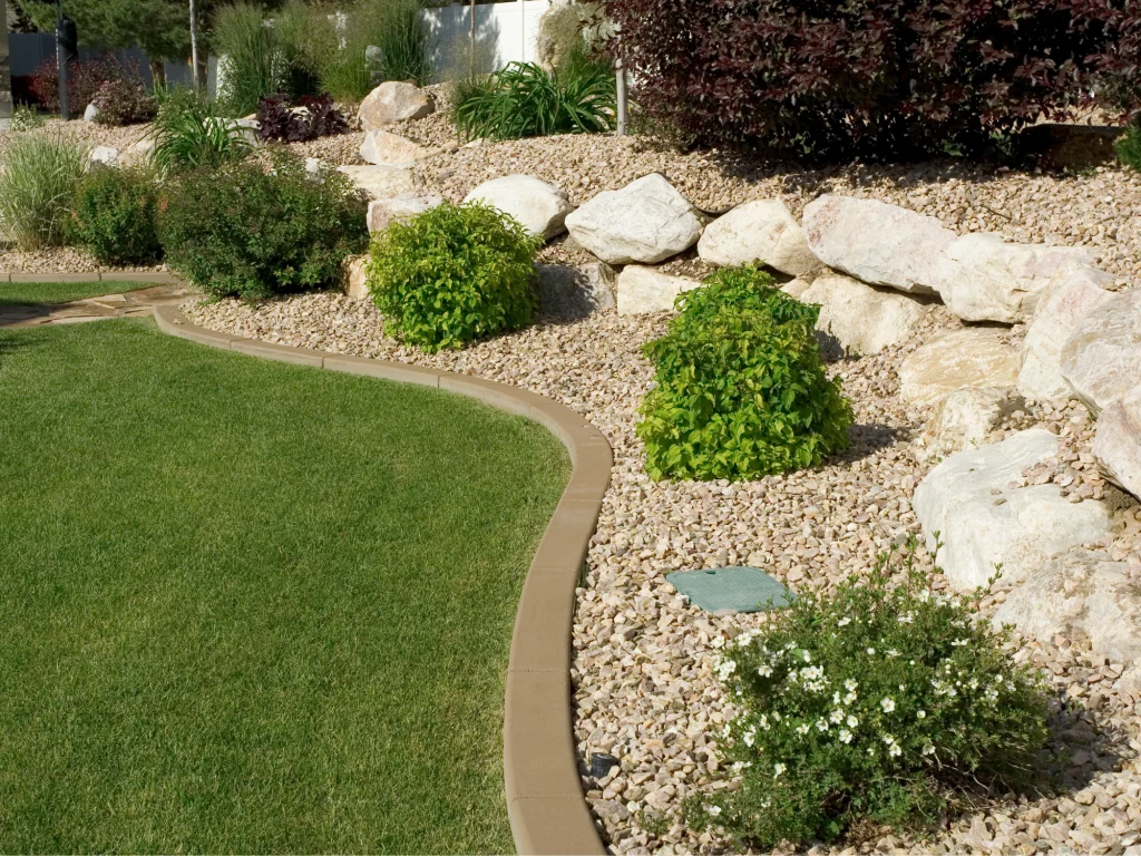 A skilled landscaper has crafted a yard featuring a curved grass border, rocks, small bushes, and plants.