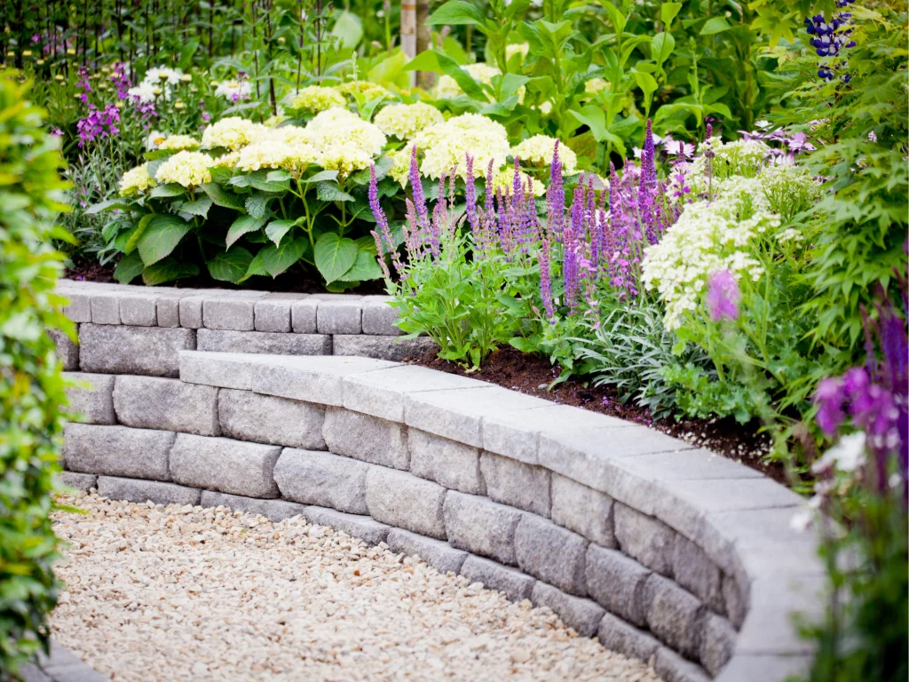 A curved stone retaining wall with steps is bordered by a garden featuring yellow, white, and purple flowers. A gravel path runs alongside the wall, making it perfect for easy yardwork in your Tacoma home.